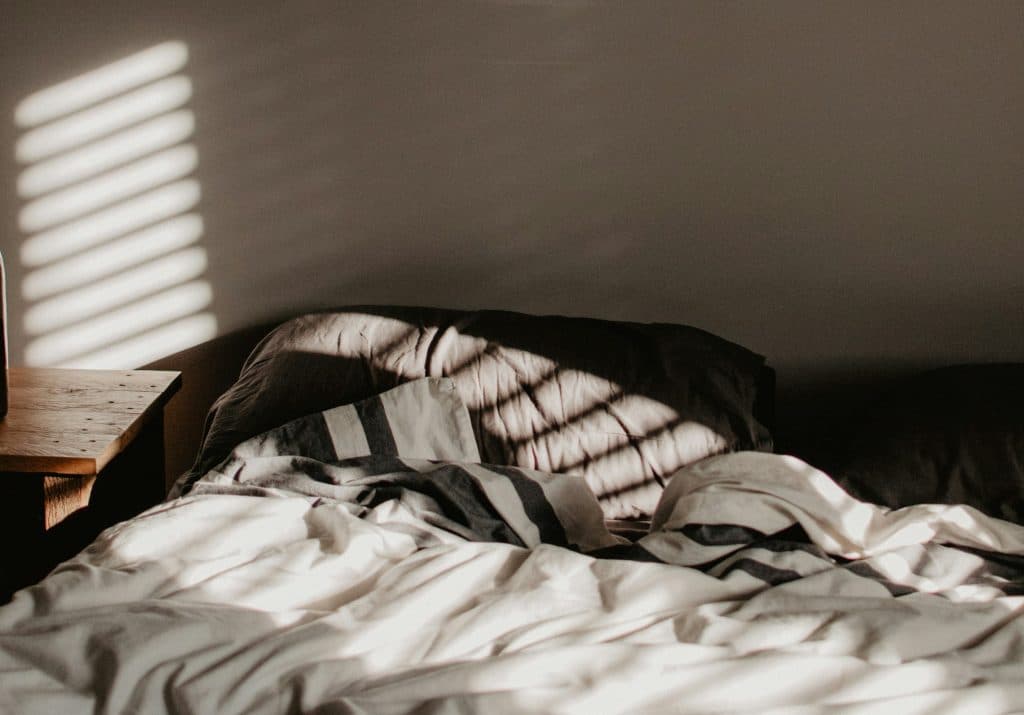 A stock photo of a bed with shadows from the blinds shining on it from the morning sun.