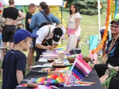 A child in a blue hat examines a table full of Pride memorabilia on a bright, sunny day.