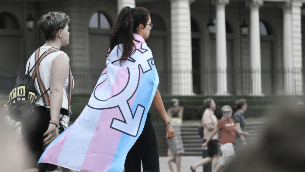 Two people walk in a desaturated photo. One person has a transgender flag around their neck that is colorized.