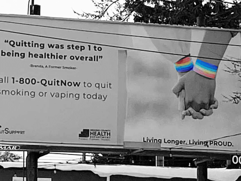 A black and white photo of a billboard with the Ohio tobacco quit line. In color are two Pride bracelets on people holding hands