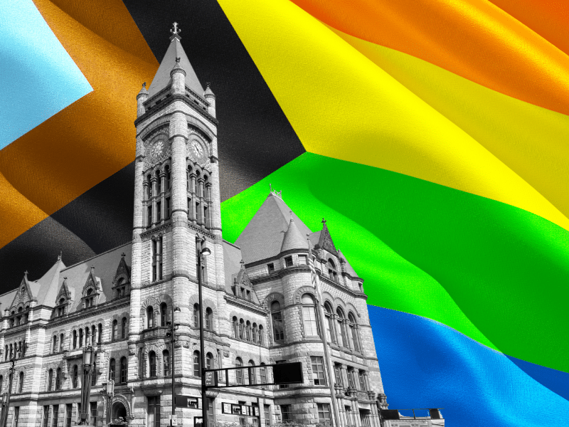 Cincinnati City Hall building is superimposed in black-and-white in front of the LGBTQ+ Progress flag.