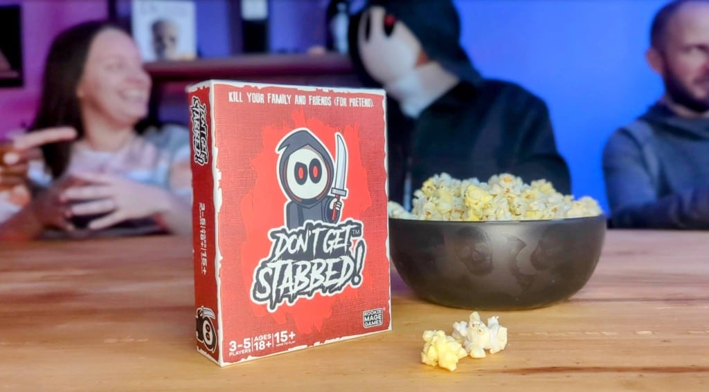 The game box of "Don't Get Stabbed" by Rookie Mage Games is on a table nearby a bowl of popcorn. Three people in the back talk about the game.